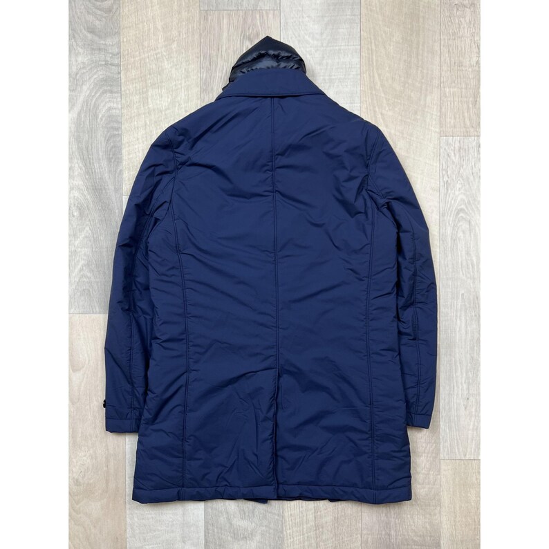 May include: A navy blue, long coat with a black hood. The coat has a button closure and a tailored fit. The back of the coat is visible, showing the seams and the overall shape. The coat is made of a smooth, possibly water-resistant material.
