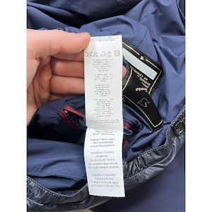 May include: A close-up of a navy blue jacket with a black label and a white tag. The tag displays washing instructions and fabric details, including 100% polyamide and duck down filling.