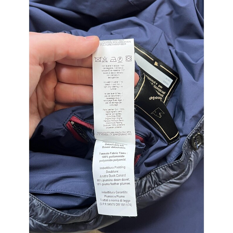May include: A close-up of a navy blue jacket with a black label and a white tag. The tag displays washing instructions and fabric details, including 100% polyamide and duck down filling.