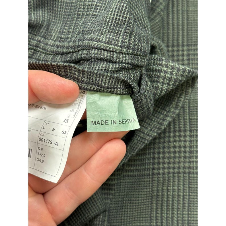 May include: A dark gray and black plaid fabric garment featuring an interior white care label with text including "MADE IN SERBIA" and "001179 -A". The textile displays a fine grid pattern with a subtle woven texture throughout the material.