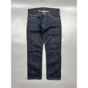 May include: A pair of dark indigo denim jeans featuring a classic five-pocket design, a button fly closure, and contrasting gold-tone stitching. The jeans have a straight-leg cut with a clean, unwashed finish and a visible brand label at the waist.