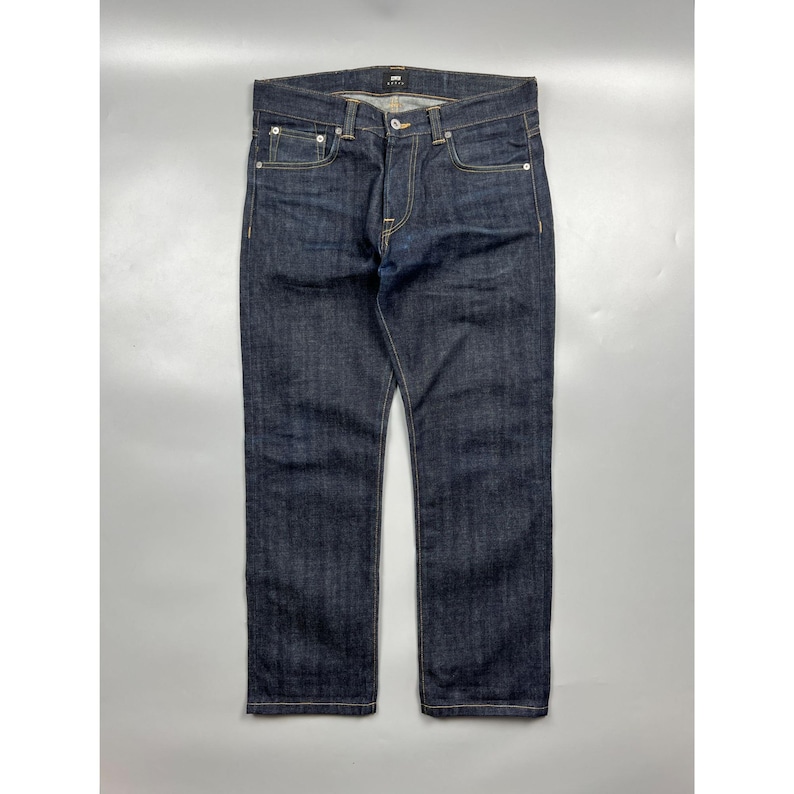 May include: A pair of dark indigo denim jeans featuring a classic five-pocket design, a button fly closure, and contrasting gold-tone stitching. The jeans have a straight-leg cut with a clean, unwashed finish and a visible brand label at the waist.