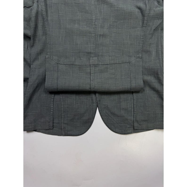 May include: A charcoal gray blazer featuring a subtle glen plaid pattern. The garment displays a structured back panel with a horizontal seam detail across the mid-section and curved hem edges at the bottom, showcasing a woven fabric texture.