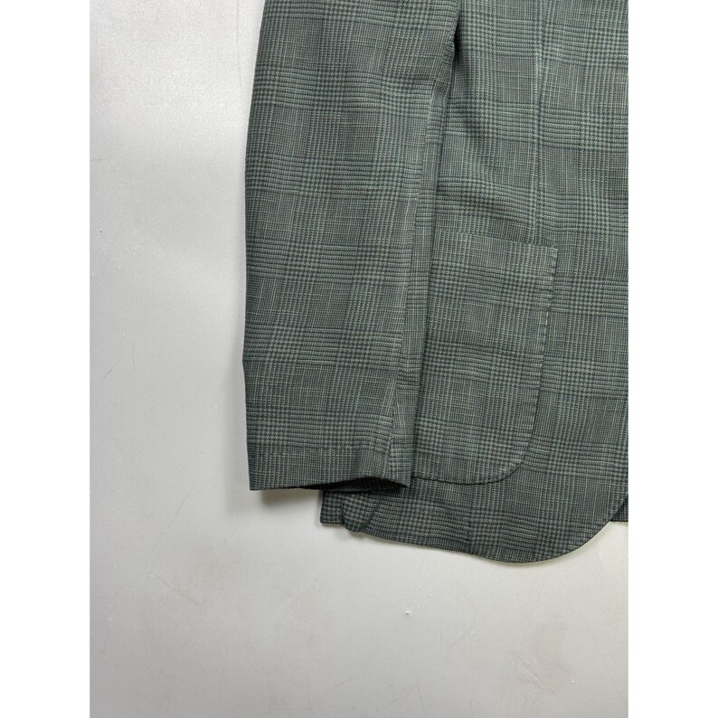 May include: A blazer in a muted olive green and dark grey glen plaid pattern. The garment features a long sleeve with a finished cuff and a single patch pocket on the lower front section. The fabric displays a fine, woven texture throughout.