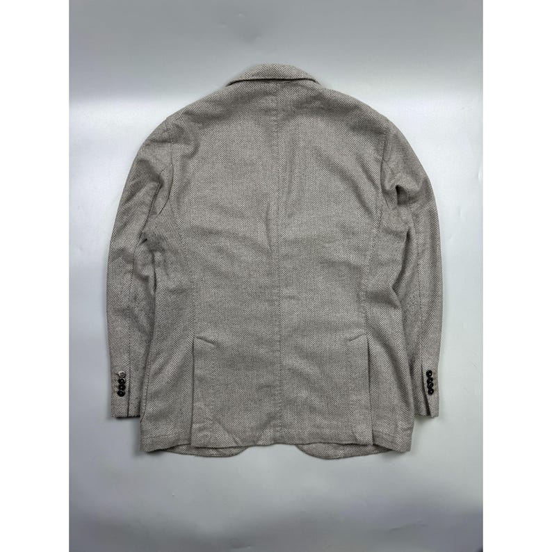 May include: A beige and brown textured blazer featuring a subtle herringbone pattern, a notched collar, long sleeves with four dark buttons on each cuff, and two vertical vents at the lower back hem, presented from a rear-facing perspective.