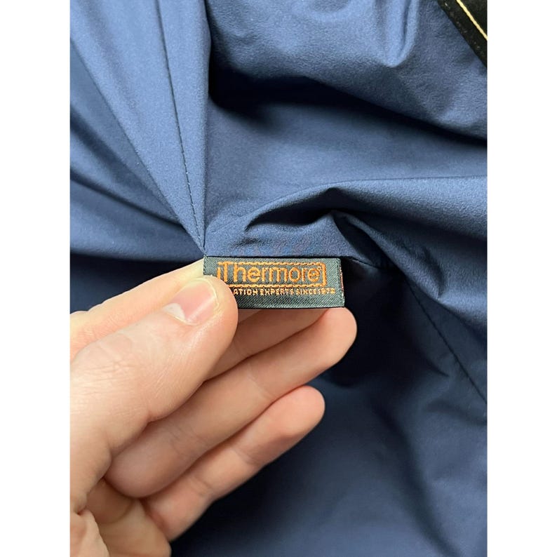 May include: Close-up of a navy blue garment with a tag that reads "Thermolo" in white text on an orange background. The tag also includes the text "INSULATION EXPERTS SINCE 1972".