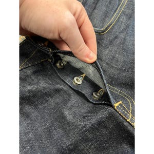 May include: Dark indigo denim jeans featuring a button fly closure with three visible silver-toned metal buttons. The fabric has a textured weave and is accented with contrasting golden-yellow stitching along the seams and button placket.