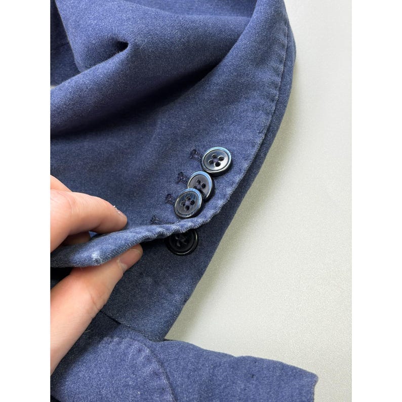 May include: A blue blazer sleeve featuring a row of four black buttons along the cuff. The fabric has a soft, matte texture with visible stitching details around the buttonholes and along the edge of the sleeve opening, showcasing the construction.