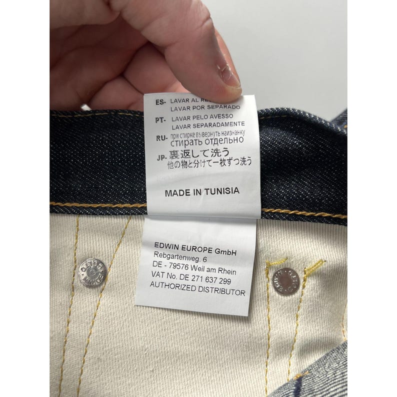 May include: A pair of dark blue denim jeans featuring a white interior care label with text in multiple languages, including instructions to wash separately and inside out. The label also states MADE IN TUNISIA and lists EDWIN EUROPE GmbH.