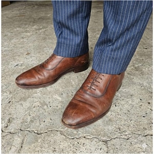 May include: A pair of brown leather dress shoes with laces. The shoes are worn and have visible creases. The shoes are paired with blue pinstripe trousers.