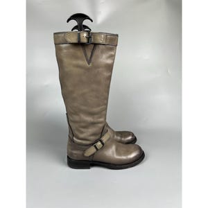 May include: A knee-high taupe leather boot featuring a rounded toe, a low block heel, and two adjustable straps with dark metal buckles positioned at the ankle and the top of the shaft. The boot includes a black plastic shoe tree insert.