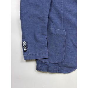 May include: A blue blazer featuring a textured fabric finish, a large patch pocket on the front, and a sleeve cuff detailed with four small, round, dark-colored buttons arranged in a vertical row with matching thread stitching.