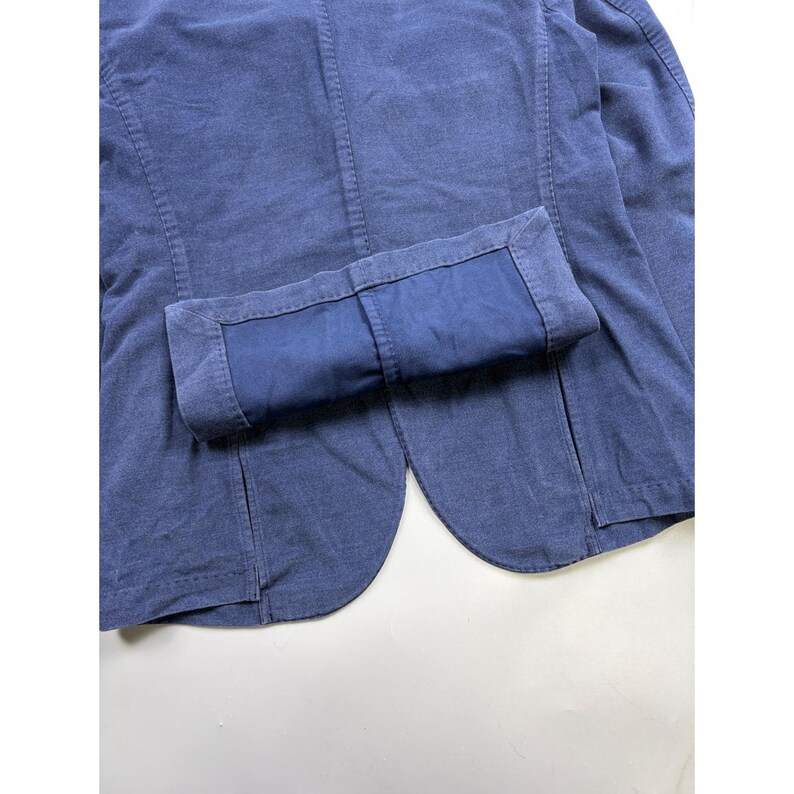 May include: A solid navy blue blazer featuring a textured fabric finish, visible vertical seam detailing, and a folded cuff on the sleeve that reveals a matching dark blue interior lining. The garment has a rounded hemline and structured stitching.