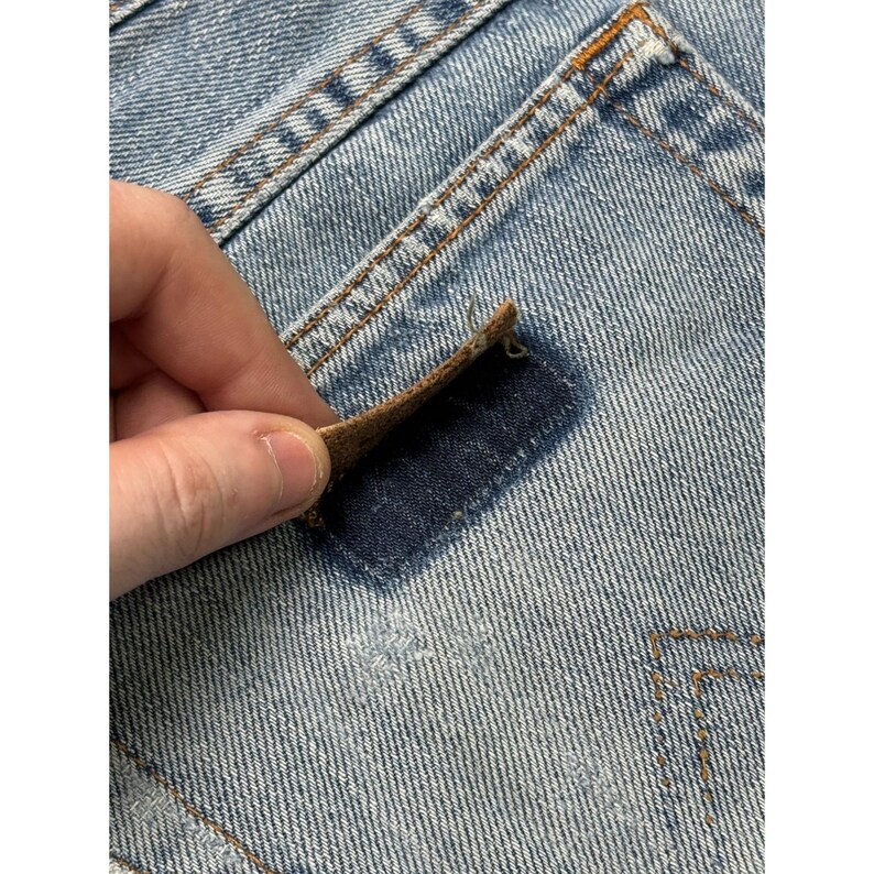 May include: Close-up of light blue denim jeans with a brown leather patch being lifted to reveal a dark blue patch underneath. The jeans have visible stitching and a distressed appearance, suggesting a vintage or worn style.