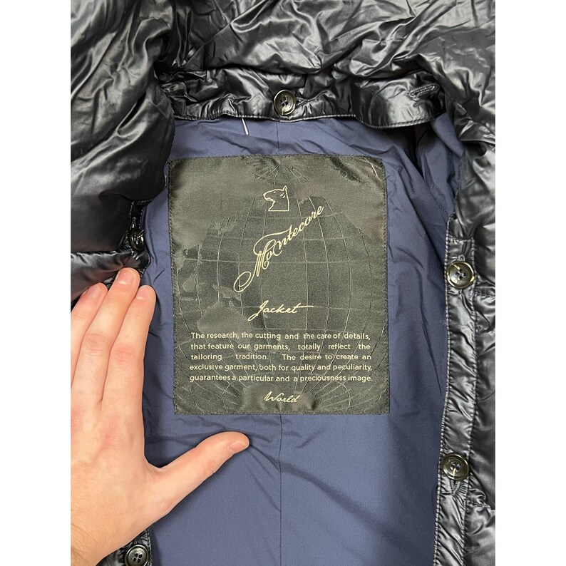 May include: Black quilted jacket with a shiny finish. The interior features a navy blue lining and a label with the brand name "Montecore" and the word "Jacket". The label also includes text about the brand's tailoring tradition.