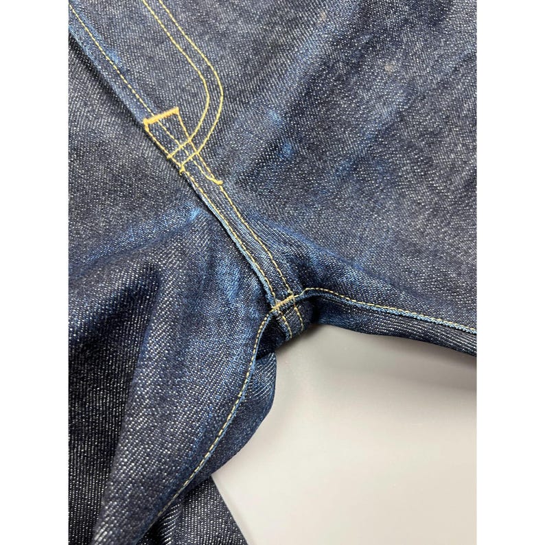 May include: Dark indigo denim jeans featuring a reinforced crotch area with visible yellow contrast stitching. The fabric displays a textured, raw denim weave with a deep blue color and sturdy construction throughout the garment seams.