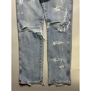 May include: Light blue denim jeans with distressed details. The jeans feature multiple rips and tears across the front of the legs, with frayed edges. The denim has a faded appearance, and the seams are visible.