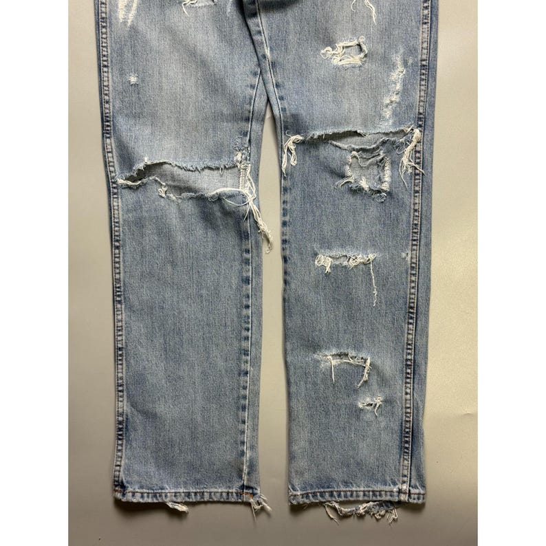 May include: Light blue denim jeans with distressed details. The jeans feature multiple rips and tears across the front of the legs, with frayed edges. The denim has a faded appearance, and the seams are visible.