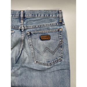May include: Light blue denim jeans with a classic five-pocket design. The back pocket features a brown leather patch with the "Wrangler" logo. The jeans have a faded wash and a straight leg cut.