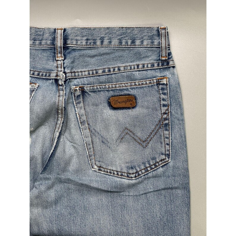 May include: Light blue denim jeans with a classic five-pocket design. The back pocket features a brown leather patch with the "Wrangler" logo. The jeans have a faded wash and a straight leg cut.