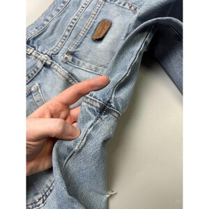 May include: Light blue denim jacket with a brown leather patch. The jacket features classic denim details like stitching and pockets. The sleeve is held by a hand, showing the jacket's texture and construction. A vintage piece.