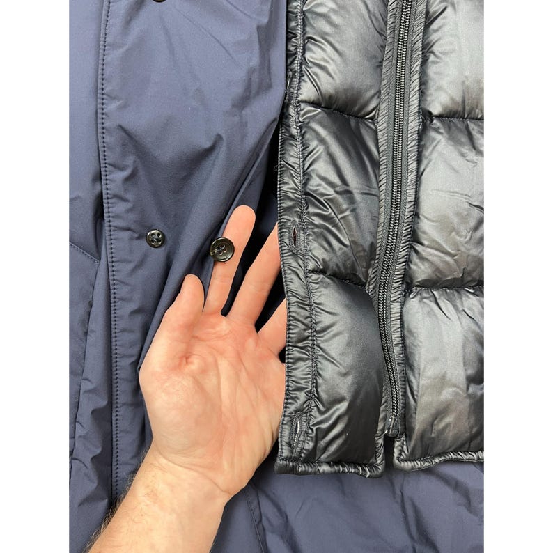 May include: A navy blue jacket with a black quilted panel and a zipper closure. The jacket has black buttons and a pocket. The interior of the jacket is visible.