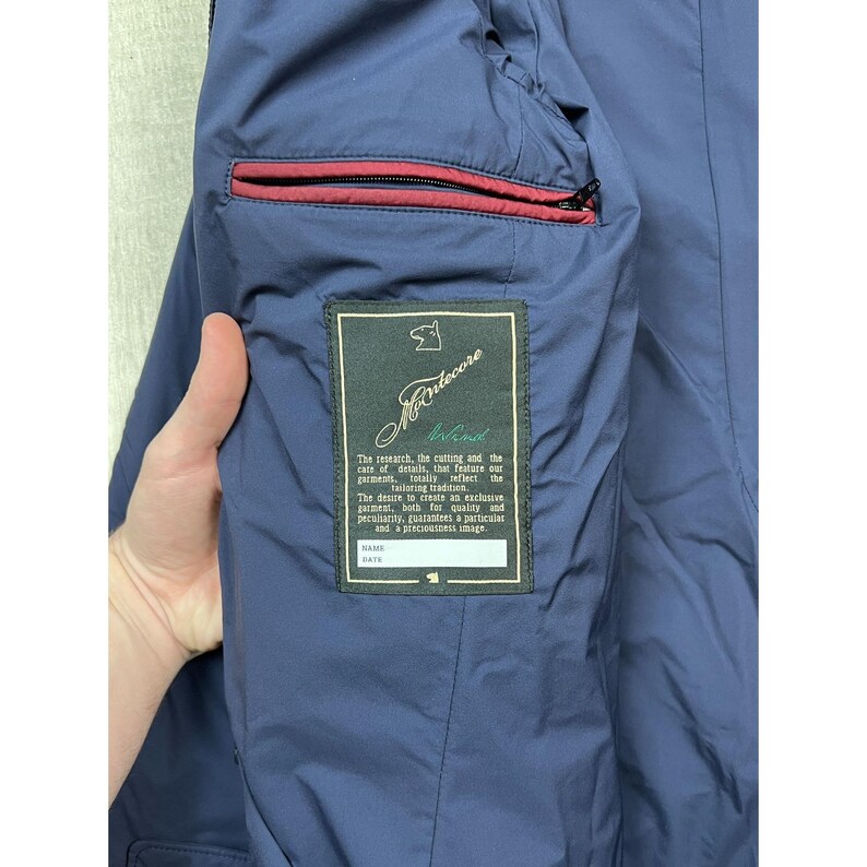 May include: Navy blue jacket interior with a red-trimmed pocket. A label with the text "Montecore" and other text is visible. The jacket appears to be made of a smooth, durable material.