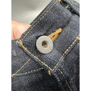 May include: Dark indigo blue denim jeans featuring a silver-toned metal button embossed with the text EDWIN. The fabric displays a visible twill weave texture with contrasting golden-yellow stitching along the waistband and button fly area.
