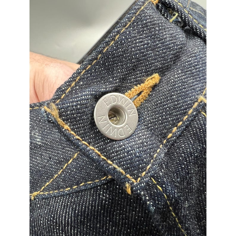 May include: Dark indigo blue denim jeans featuring a silver-toned metal button embossed with the text EDWIN. The fabric displays a visible twill weave texture with contrasting golden-yellow stitching along the waistband and button fly area.