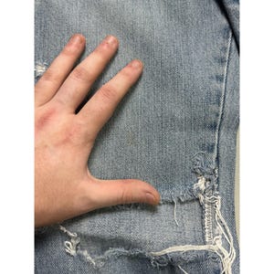 May include: Light blue denim jeans with distressed details, including rips and frayed edges. The jeans are shown with a hand resting on the fabric, highlighting the texture and wear.