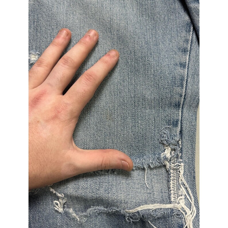 May include: Light blue denim jeans with distressed details, including rips and frayed edges. The jeans are shown with a hand resting on the fabric, highlighting the texture and wear.