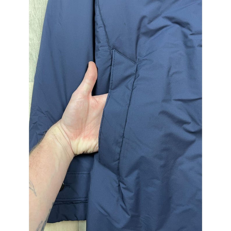 May include: Close-up of a navy blue jacket with a hand in the pocket. The jacket has a smooth, matte finish and a simple, classic design. The pocket is on the front of the jacket.