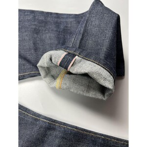 May include: Dark indigo denim jeans featuring a cuffed hem that reveals a white interior with a distinct red and white selvedge ID line. The fabric has a visible twill texture, and the seams are reinforced with contrasting gold-colored stitching.