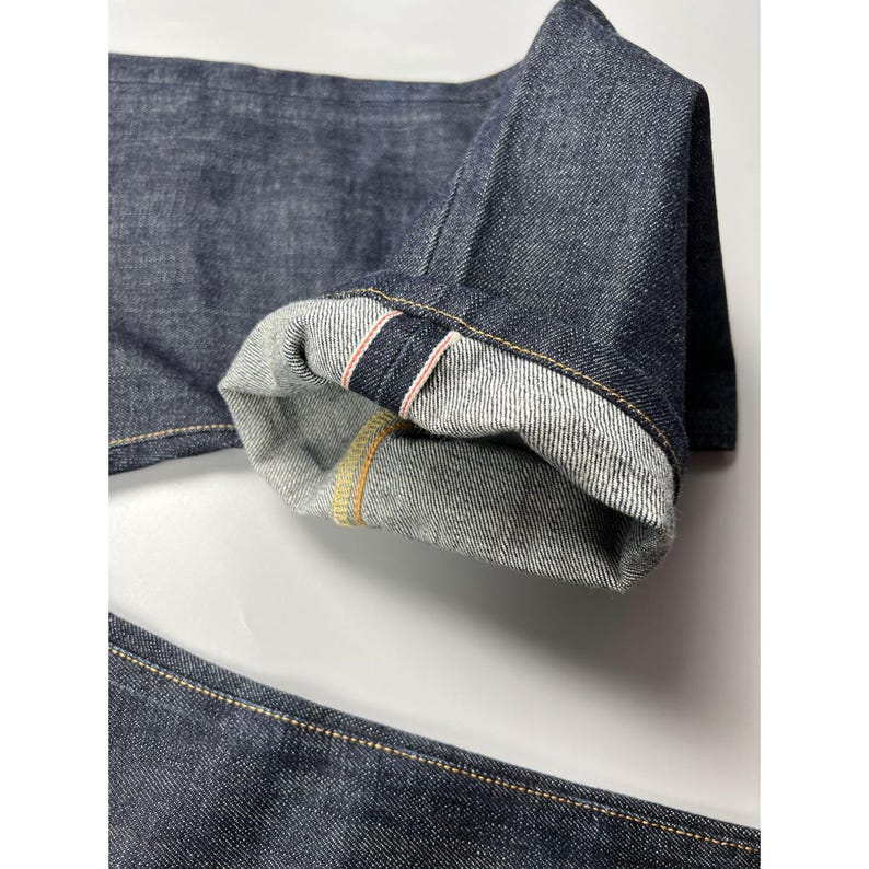 May include: Dark indigo denim jeans featuring a cuffed hem that reveals a white interior with a distinct red and white selvedge ID line. The fabric has a visible twill texture, and the seams are reinforced with contrasting gold-colored stitching.