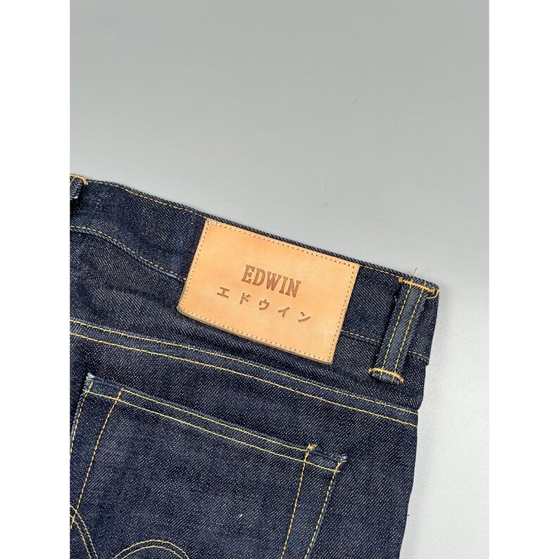 May include: Dark indigo denim jeans featuring a tan leather patch on the back waistband. The patch is embossed with the text EDWIN and the Japanese characters エドウイン. The jeans are constructed with visible gold-toned stitching along the seams.
