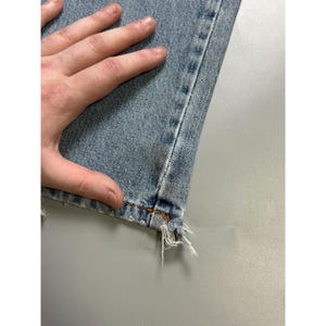 May include: Close-up of light blue denim jeans with a frayed hem. The jeans have a classic five-pocket design and are made of a durable denim fabric. The stitching is visible along the seams and the hem is distressed.