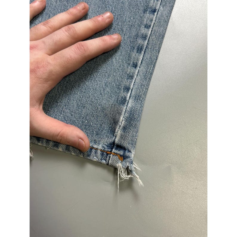 May include: Close-up of light blue denim jeans with a frayed hem. The jeans have a classic five-pocket design and are made of a durable denim fabric. The stitching is visible along the seams and the hem is distressed.