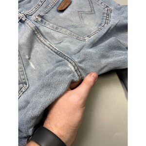 May include: Light blue denim jeans with a distressed look. The jeans feature a classic five-pocket design, a brown leather patch with a logo, and a faded wash. The jeans are held by a hand, showcasing the texture and details of the fabric.