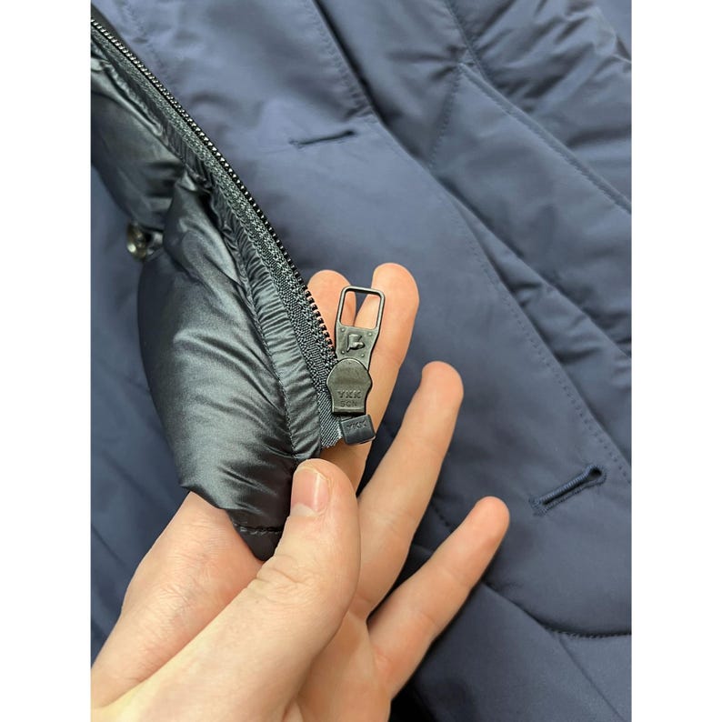 May include: Close-up of a navy blue quilted jacket with a black zipper. The zipper is open, revealing the interior lining. The jacket has a button closure and a smooth, matte finish.