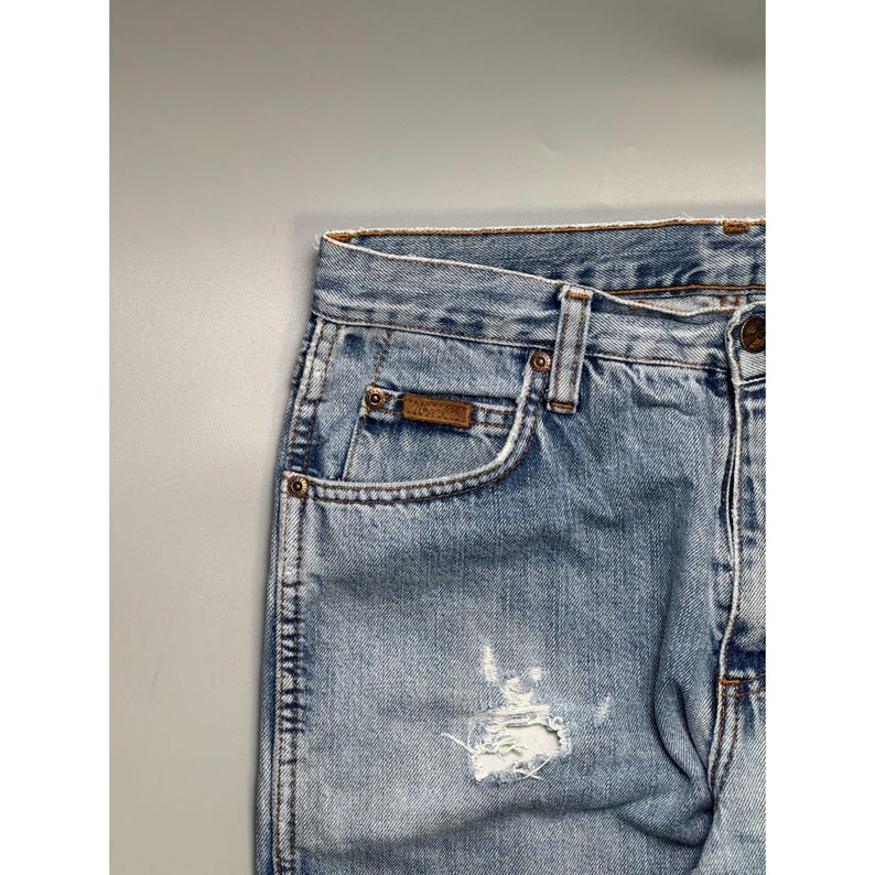 May include: Light blue denim jeans with a distressed, worn look. Features a small brown leather patch on the front pocket and a ripped section on the leg. Classic five-pocket design with metal rivets and a button closure.