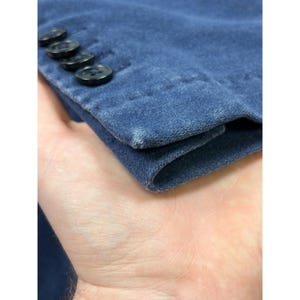 May include: A blue suede sleeve cuff featuring four black buttons aligned in a row along the edge. The fabric displays a soft, matte texture with slight variations in color intensity and a small area of lighter wear on the corner edge.