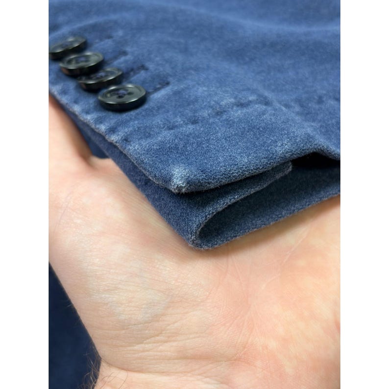 May include: A blue suede sleeve cuff featuring four black buttons aligned in a row along the edge. The fabric displays a soft, matte texture with slight variations in color intensity and a small area of lighter wear on the corner edge.