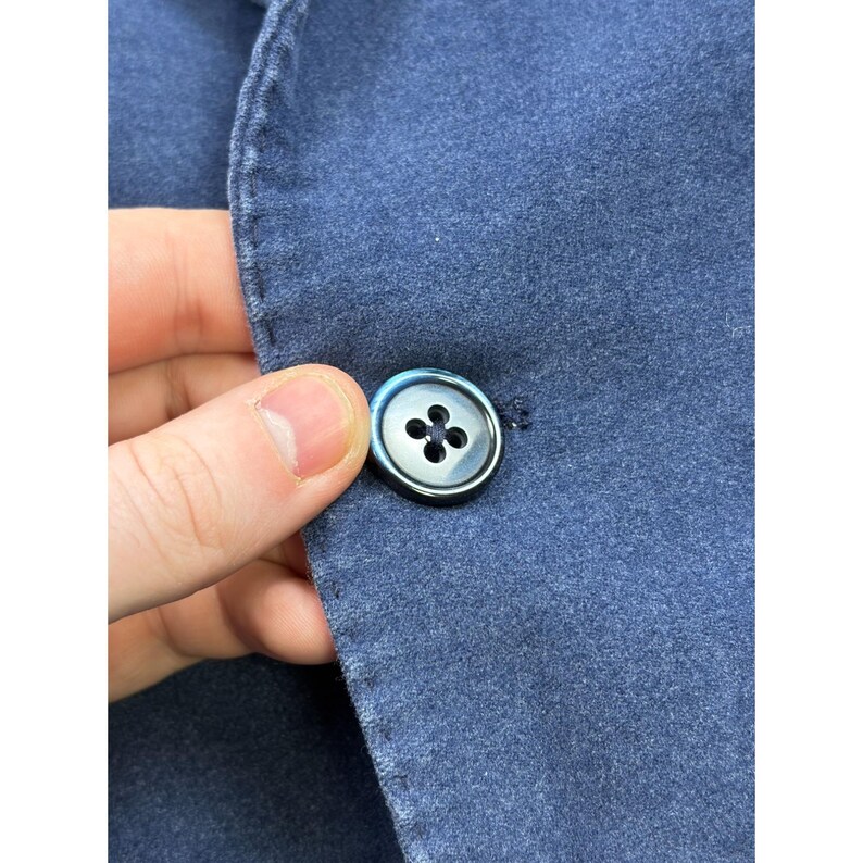 May include: A blue blazer featuring a textured fabric surface and a single dark, iridescent four-hole button. The garment displays visible stitching along the lapel edge, highlighting the construction of the blue textile material.