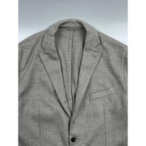 May include: A light gray and white blazer featuring a classic herringbone pattern. The garment includes a notched lapel, a single visible dark button closure at the front, and a single welt pocket on the left side of the chest area.
