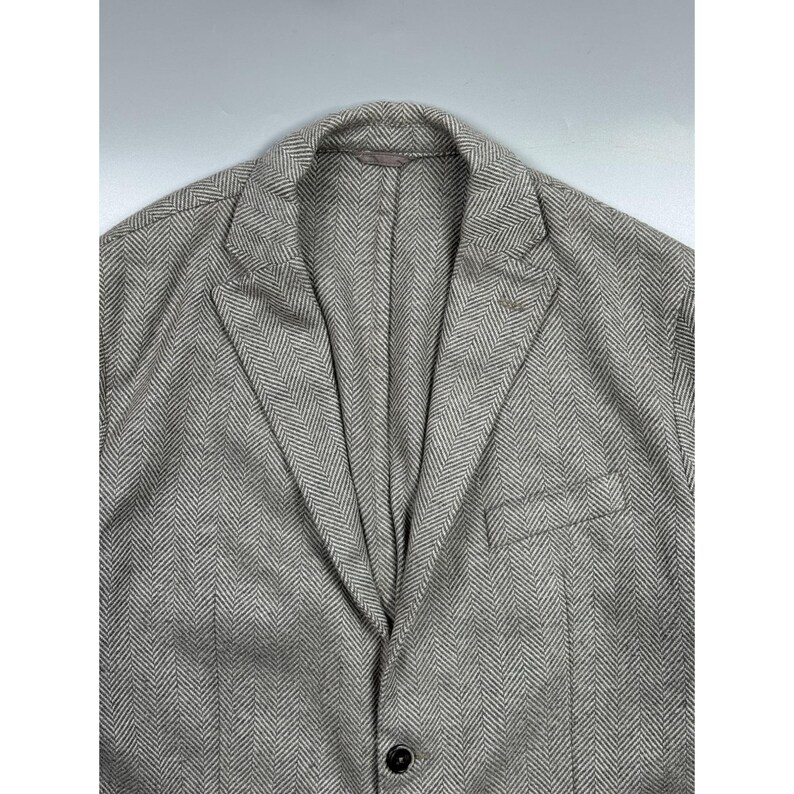 May include: A light gray and white blazer featuring a classic herringbone pattern. The garment includes a notched lapel, a single visible dark button closure at the front, and a single welt pocket on the left side of the chest area.