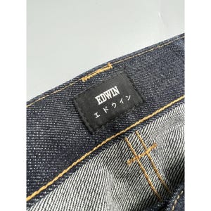 May include: Dark indigo denim jeans featuring a black rectangular fabric label stitched onto the waistband. The label displays the text EDWIN in white capital letters above the Japanese katakana characters for Edwin. Gold contrast stitching is visible.