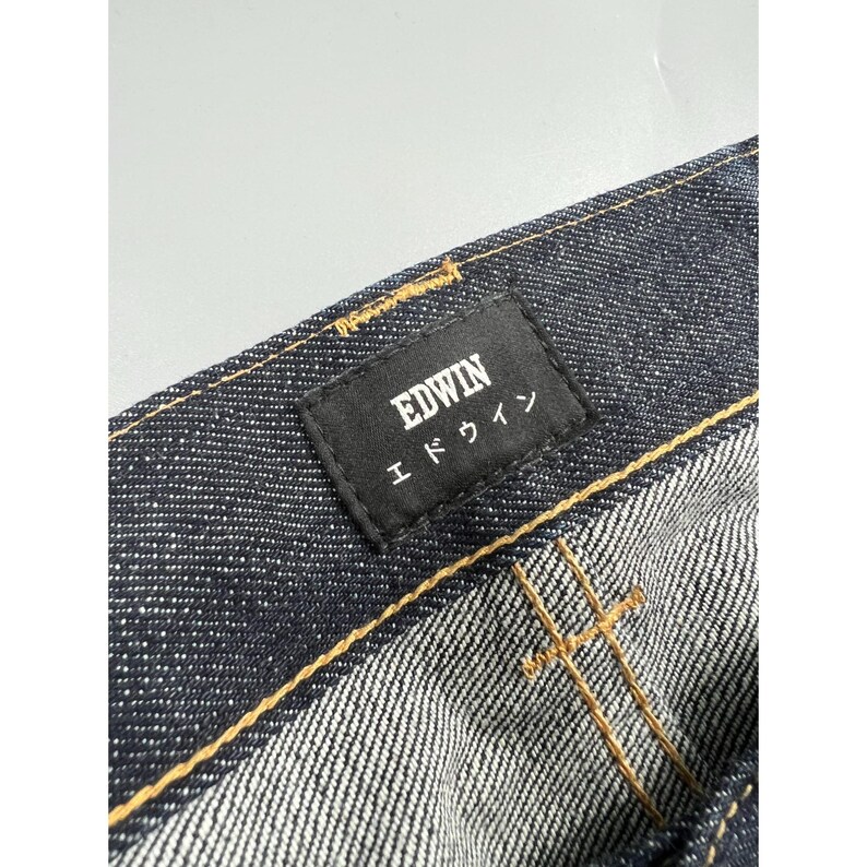 May include: Dark indigo denim jeans featuring a black rectangular fabric label stitched onto the waistband. The label displays the text EDWIN in white capital letters above the Japanese katakana characters for Edwin. Gold contrast stitching is visible.