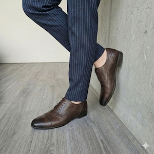 May include: Brown leather dress shoes with a classic lace-up design. The shoes have a polished finish and a low heel. The image shows the shoes paired with navy blue pinstripe trousers.