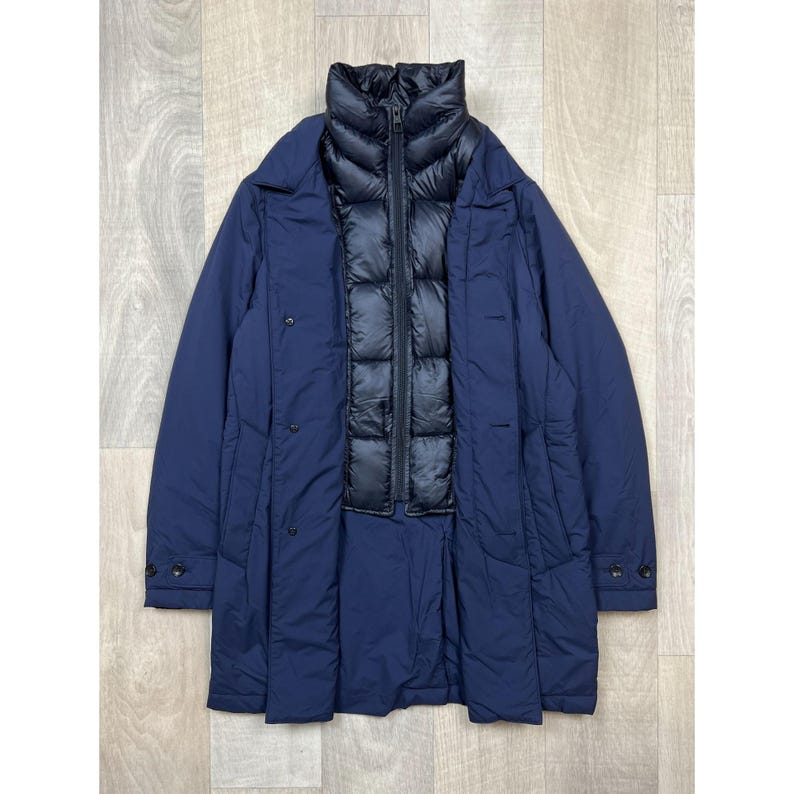 May include: A navy blue coat with a black quilted inner layer. The coat has a high collar, button closures, and a long, streamlined design. The outer layer is a smooth, solid color.