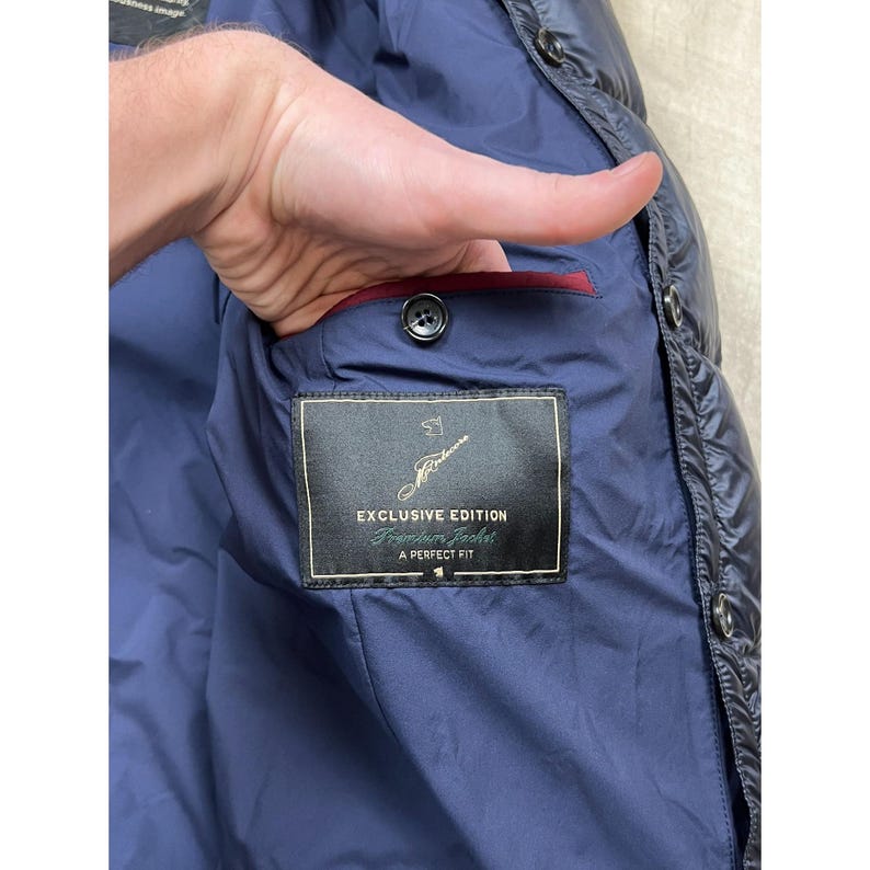 May include: Navy blue quilted jacket with a black button closure and a red interior lining. The jacket features a label that reads "EXCLUSIVE EDITION Traveler Jacket A PERFECT FIT".